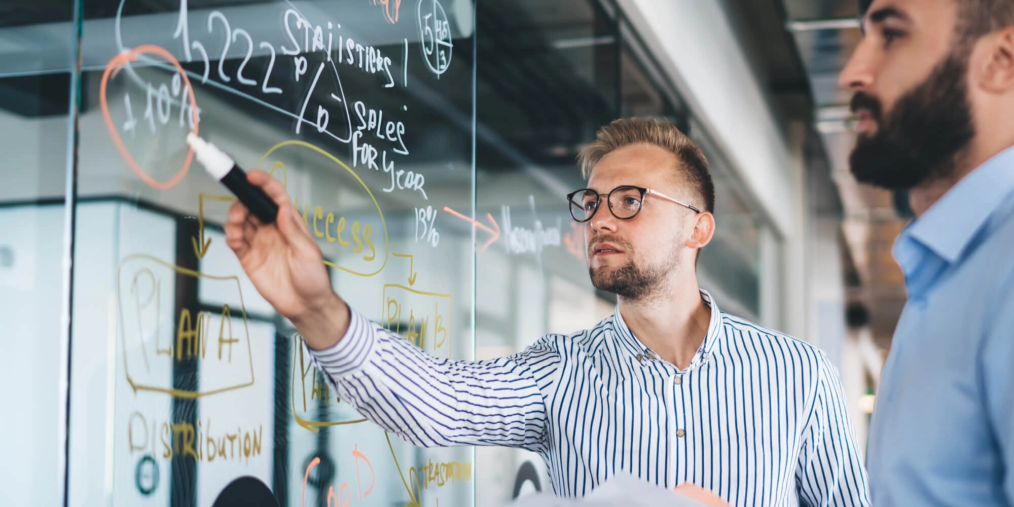 Two men collaboratively writing on a glass whiteboard, engaged in a discussion or brainstorming session
