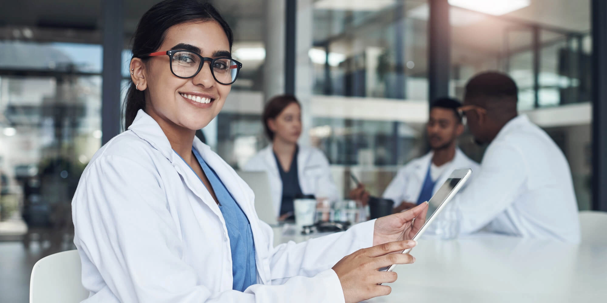 A woman in a lab gown with an Ipad smiling for the camera