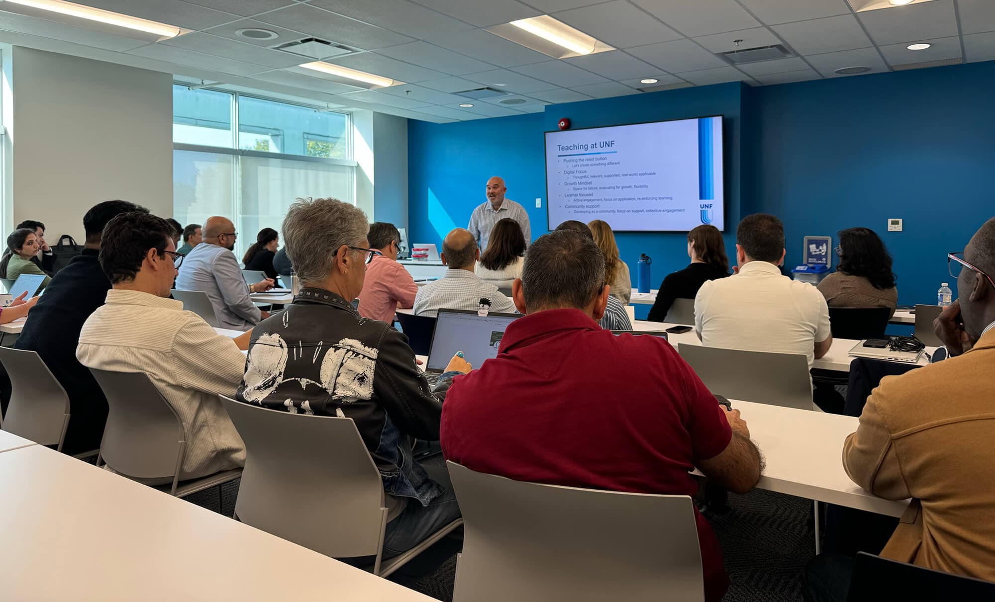 A man stands at the front, presenting to an engaged audience of diverse individuals seated in a conference room