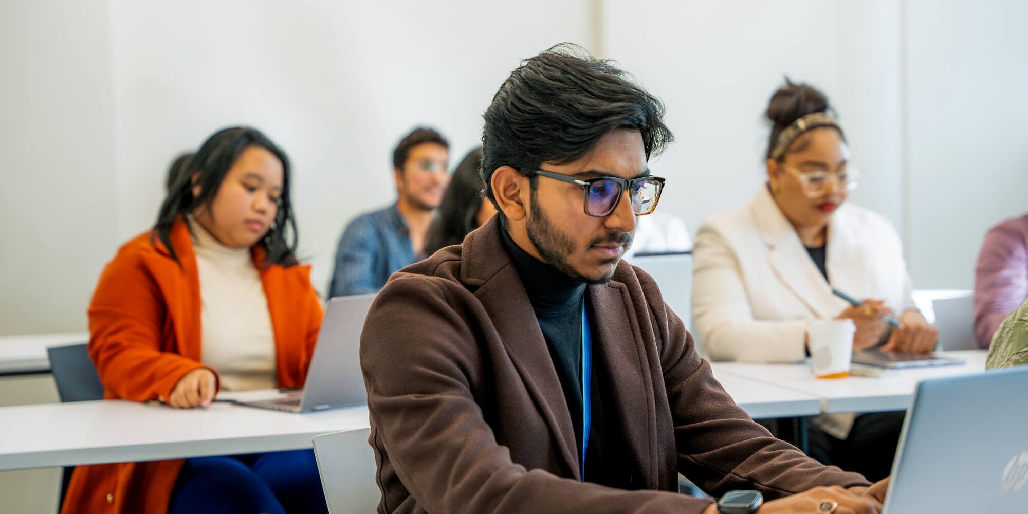 A man wearing glasses is focused on his laptop, engaged in work at a desk