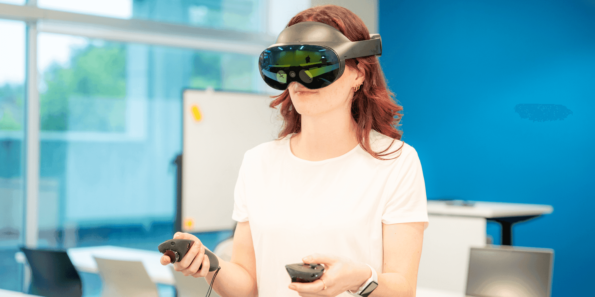 A woman wearing a VR headset and holding its controllers on a classroom setting