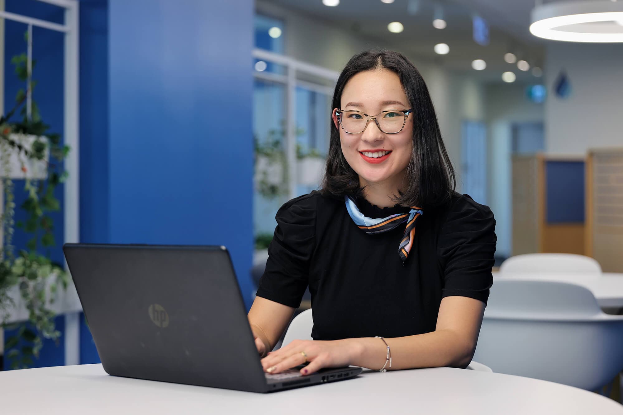 A female student with glasses and a laptop sitting inside UNF campus looking at the camera