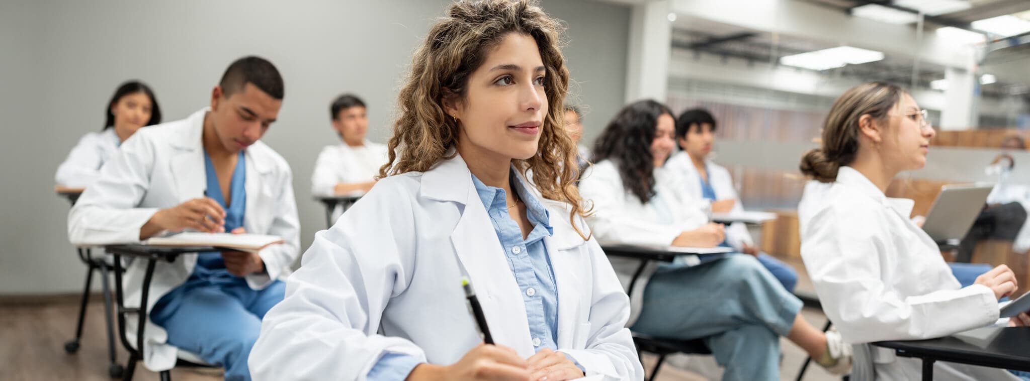 A woman in a lab coat is seated at a desk, engaged in her work in a classroom setting