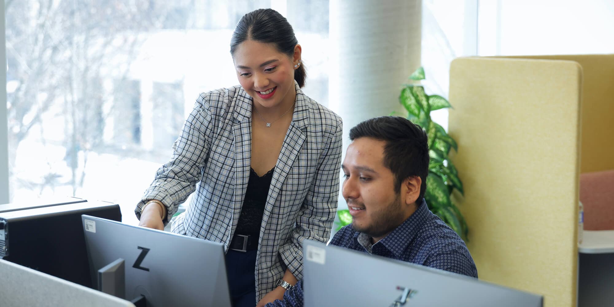 A woman standing and a man sitting in front of a desktop computer discussing something