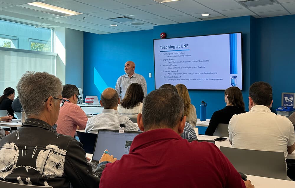 A man stands at the front, presenting to an engaged audience of diverse individuals seated in a conference room