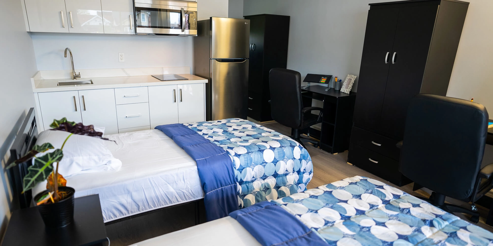 A small bedroom featuring two beds, two desks, and a refrigerator, creating a cozy and functional living space