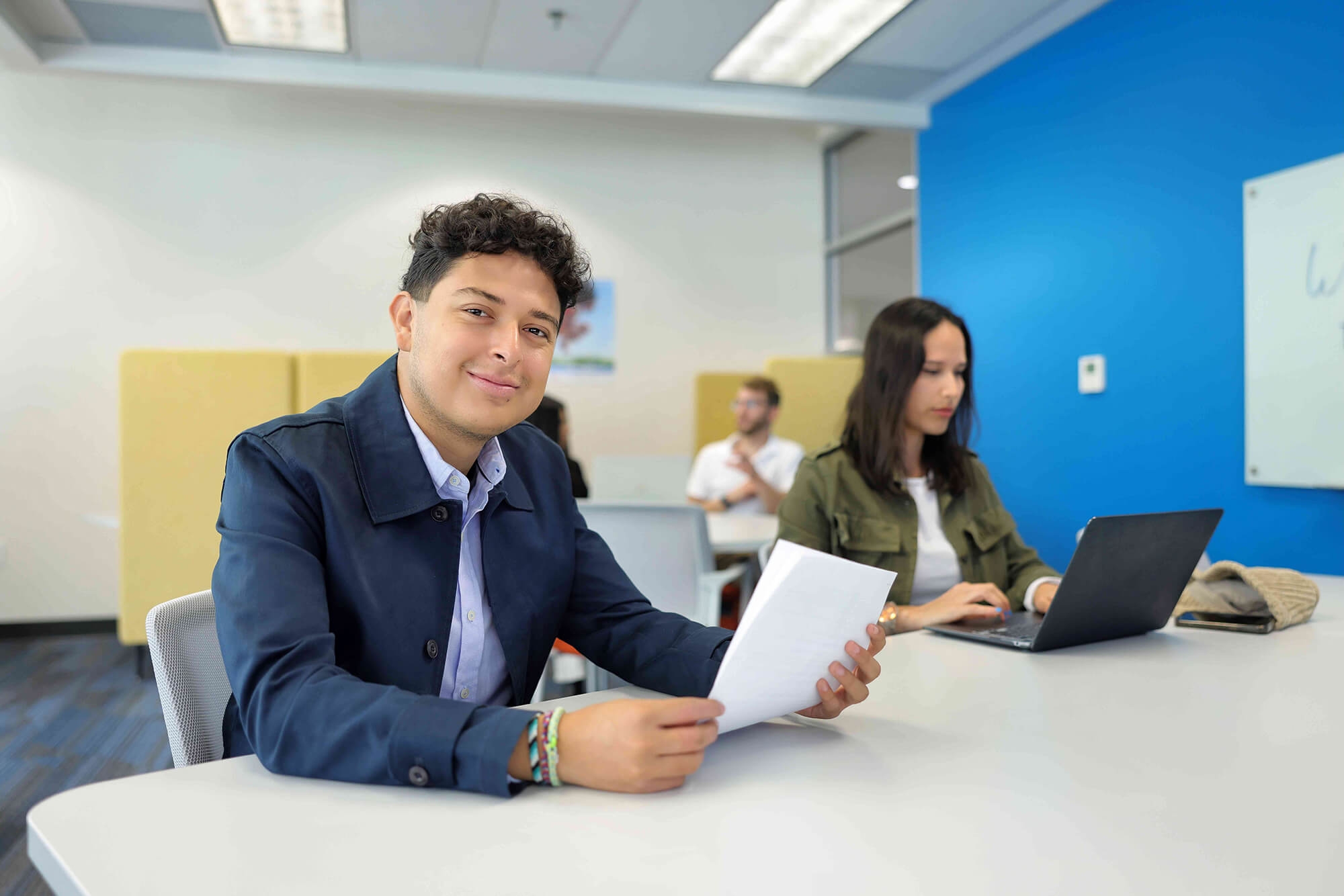 A guy holding a paper with a girl in front of a laptop both sitting on a table on a comfortable environment