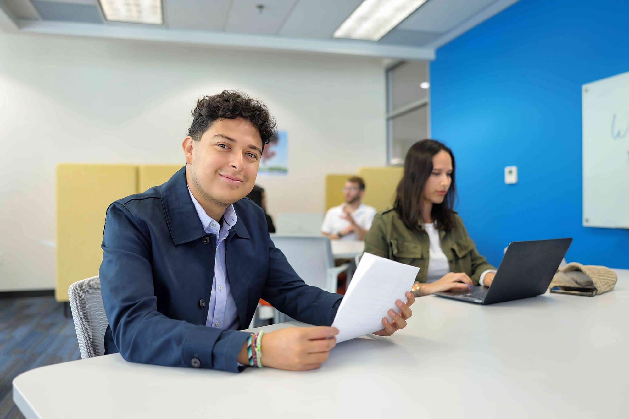 A guy holding a paper with a girl in front of a laptop both sitting on a table on a comfortable environment
