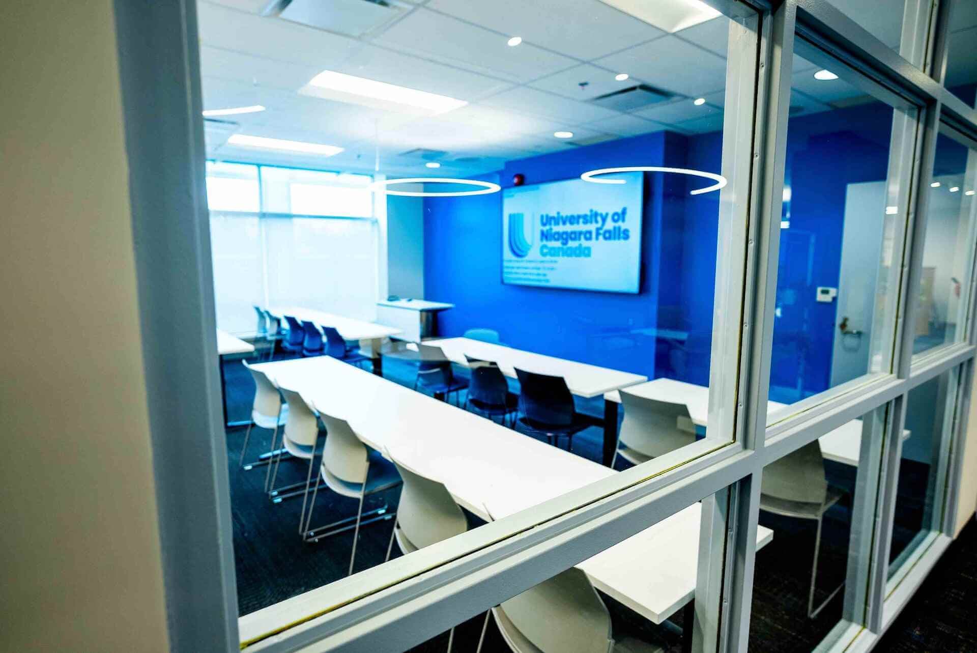 A view of a classroom featuring white tables and chairs from outside a window