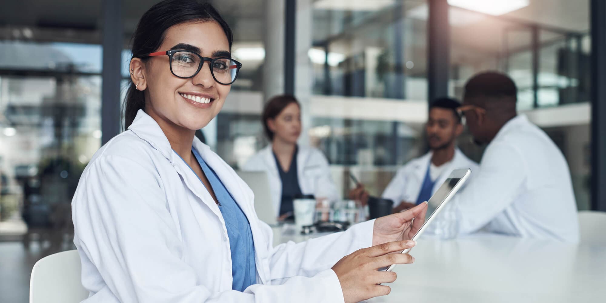 A woman in a lab gown with an Ipad smiling for the camera